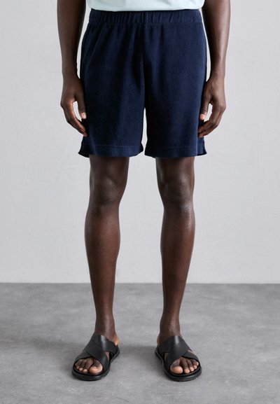 Dunkelblaue Frottee-Shorts mit einem elastischen Bund und umgeschlagenen Säumen; kombiniert mit schwarzen Riemchensandalen.