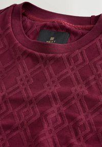 Weinrote Sweatshirt mit texturiertem geometrischen Muster, Rundhalsausschnitt und sichtbarem Markenlabel im inneren Halsbereich.