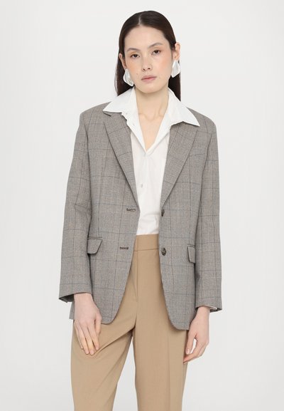 Blazer gri în carouri, cu guler ascuțit, două butoane frontale și buzunare aplicate, purtat cu o cămașă albă și pantaloni bej. Textură netedă.