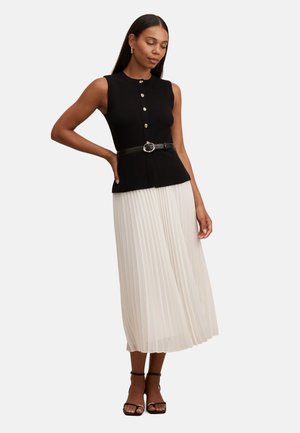 Femme portant un top noir sans manches avec des boutons et une ceinture, associé à une jupe midi plissée blanche et des sandales noires à talons, debout avec une main sur la hanche.