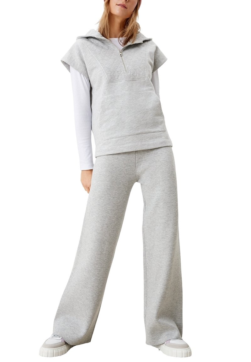 Femme portant un sweat à capuche sans manches gris clair à fermeture éclair quart de zip par-dessus une chemise blanche à manches longues, un pantalon de survêtement gris à jambes larges et des baskets blanches.