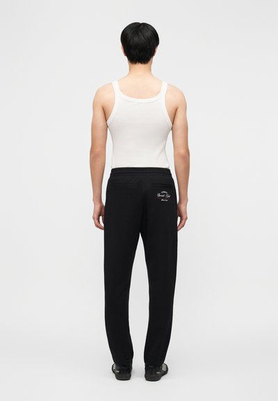 Άνδρας που βλέπει προς τα πίσω φοράει λευκό αμάνικο, μαύρη φόρμα με κείμενο στην πίσω τσέπη και μαύρα παπούτσια σε απλό λευκό φόντο.