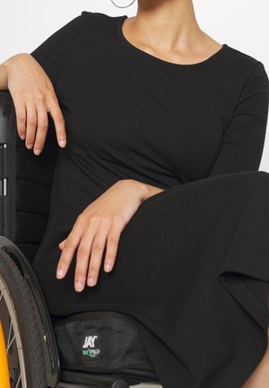 Osoba ubrana w czarną sukienkę siedząca na wózku inwalidzkim, jedną ręką opierająca się na poręczy wózka, a drugą na kolanach.