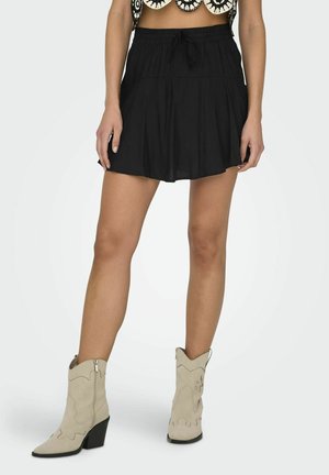 Femme portant une jupe noire courte avec une taille à cordon et des bottes de cowboy beige sur fond blanc.
