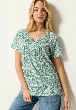 Femme blonde portant un chemisier à manches courtes vert et blanc à motifs avec des boutons et des plis, associé à un jean bleu, debout contre un mur blanc.