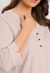 Frau mit langen braunen Haaren, die ein hellrosa V-Ausschnitt-Oberteil mit drei schwarzen Knöpfen und einem Schlitz am Ärmel trägt und den Kragen mit der Hand hält.