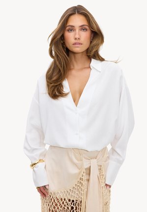 Femme aux longs cheveux bruns portant un chemisier blanc ample et une jupe beige avec des détails macramé noués à la taille, debout devant un fond uni.