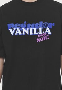 Juodas medvilnės marškinėliai su ryškiu, spalvingu užrašu: "pegador VANILLA Super Soft!" mėlyna, balta ir rožine spalvomis, naudojant žaismingą šriftą.