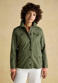 Moteris su garbanotais plaukais, vilkinti olive žalios spalvos užsegamą švarką ir baltas kelnes priešais paprastą smėlio spalvos foną.