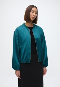 Tűzkék selyem bomber dzseki hosszú ujjakkal és elasztikus aljjal, elülső cipzárral és oldalsó zsebekkel, fekete felsőre rétegezve.