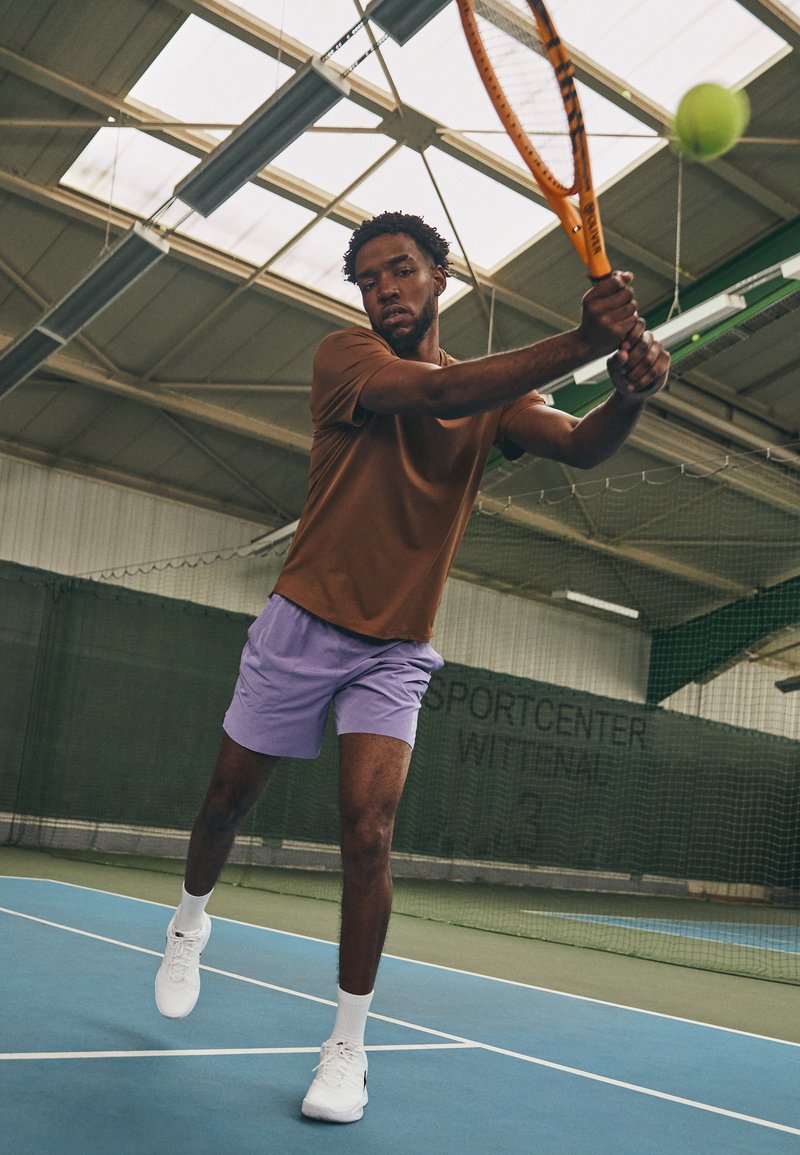 Ein männlicher Athlet trägt ein braunes kurzärmliges Shirt und lila Sportshorts, während er auf einem Hallentennisplatz einen Tennisschläger schwingt. Weiße Turnschuhe sind sichtbar.