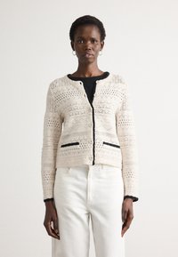 Cardigan a maglia beige con motivi geometrici, rifiniture nere lungo i bordi e due tasche nere. Bottoni sulla parte anteriore.