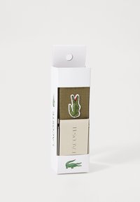 Olive zöld szövetből készült Lacoste öv, fehér és zöld krokodil logóval. A csomagolás fehér, dombornyomott márkanevével és akasztási lehetőséggel rendelkezik.
