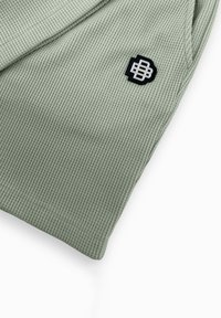 Lichtgroene, textuurstof met een ribbelpatroon; voorzien van een zwart en zilver logo-embleem. Focus op vorm en constructiedetails.