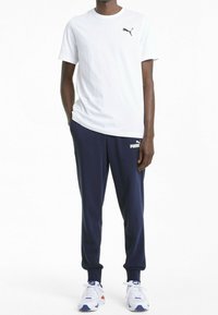 Vit kortärmad T-shirt med en svart Puma-logotyp, kombinerad med marinblå sweatpants med en vit Puma-logotyp, och vita träningsskor med accenter.