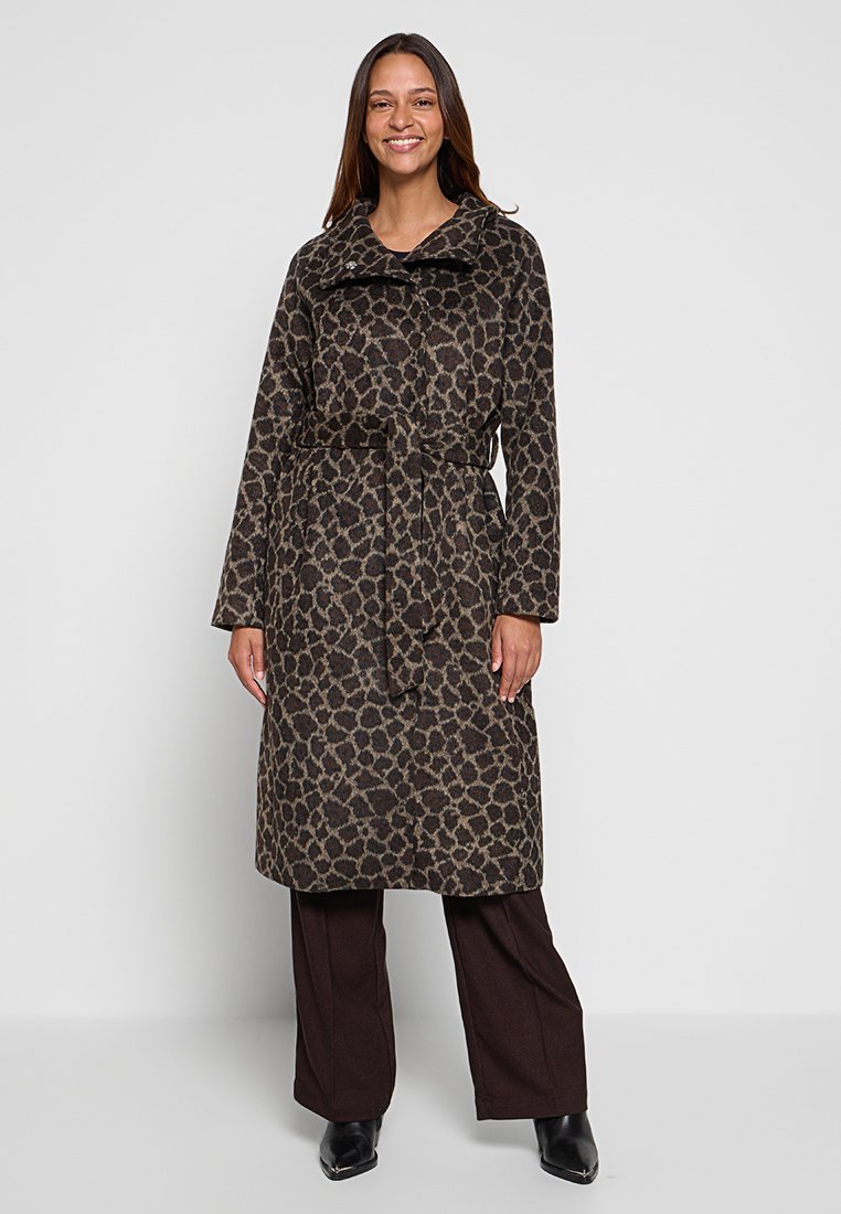 Donna sorridente che indossa un cappotto a pois leopardati con cintura sopra pantaloni marrone scuro e scarpe nere con punta, in piedi contro uno sfondo semplice.