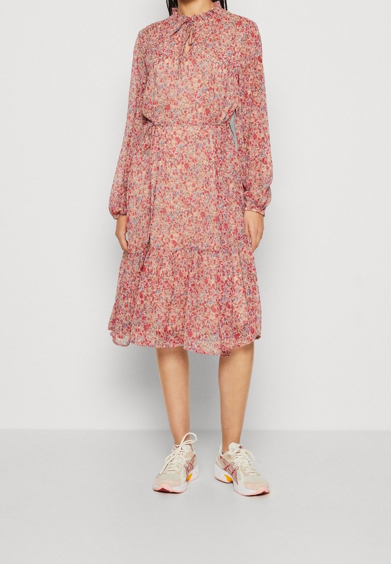 Robe florale légère en rose doux avec des accents bleus, manches longues, encolure froncée avec lien, et ourlet volanté, associée à des baskets beiges.