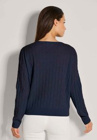 Frau mit langen dunklen Haaren, die einen marineblauen, kabelstrickartigen Pullover und weiße Hose trägt, mit dem Rücken zu einem schlichten hellen Hintergrund.