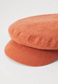 Căciulă plată dintr-un material moale, portocalie, cu un vârf rotund, margine lată și detalii de cusătură vizibile de-a lungul marginii.