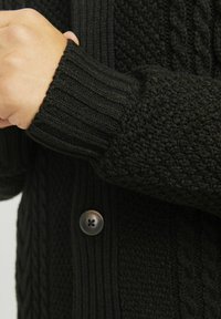 Crni pleteni kardigan s kabelastom teksturom, rebrastim manžetama i okruglim smeškim dugmetom. Mekana, topla tkanina s raznolikim uzorcima šivanja.
