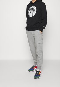 Osoba w czarnej bluzie Puma, szarych spodniach dresowych Puma i wielokolorowych trampkach stoi z rękami w kieszeniach na białym tle.