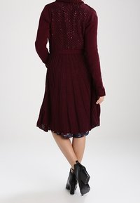 Burgunderfarbene Strickjacke mit einem strukturierten, Spitzenrücken und einem plissierten Rock. Sie hat einen hohen Kragen und eine taillierte Passform, kombiniert mit schwarzen, hochhackigen Stiefeln.