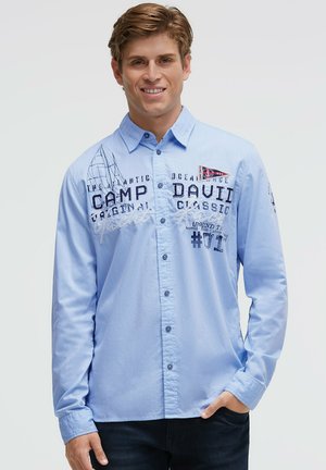 Junger Mann lächelnd, trägt ein hellblaues Hemd mit Knöpfen, dunklem Text und kleinem dreieckigem Flaggen-Design auf der Brust.