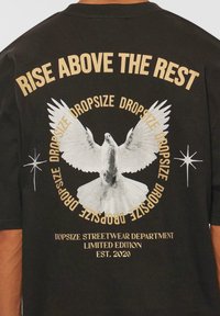 Sort bomulds T-shirt med et stort grafik af en hvid due, omgivet af tekst i guld. Teksten inkluderer "Rise Above The Rest" og mærkets oplysninger.