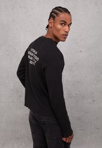 Maglietta nera a maniche lunghe con collo a barca, con testo bianco sul retro e orlo grezzo. Abbinata a jeans scuri.