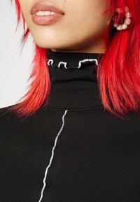Svart ribbad polotröja med volangkrage och vit sömnadsdetalj. Modellen har livligt röd hår och synliga hoop-örhängen.