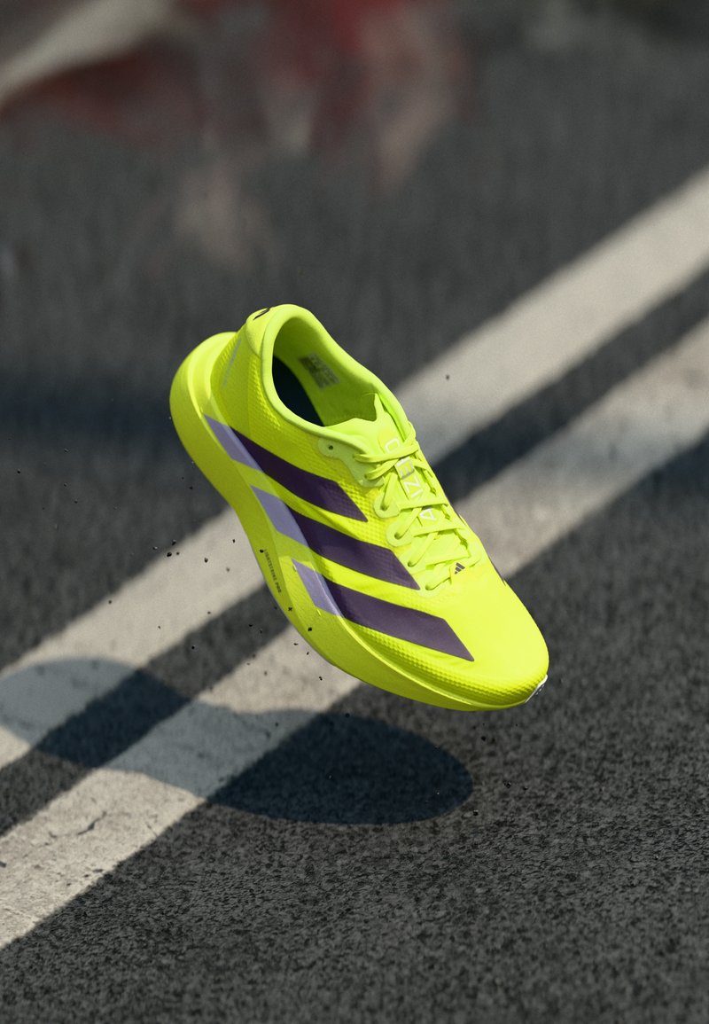 Neon-gelber Sportschuh mit violetten und weißen Streifen, leichtem Mesh-Obermaterial, gepolsterter Sohle, stromlinienförmigem Design und flachen Schnürsenkeln.