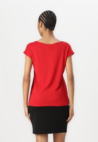 Kvinna med flätat hår står vänd bort, klädd i en röd kortärmad topp och en svart kjol framför en vit bakgrund.