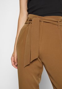 Pantalones de tiro alto marrones hechos de una tela suave. Presentan un detalle de cinturón con lazo y pliegues, con un diseño de pierna recta y textura suave.