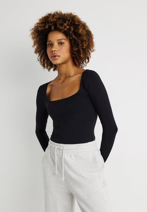 Mladá žena s kučeravými vlasmi, oblečená v čiernom tričku s dlhými rukávmi a štvorcovým výstrihom a svetlošedých nohaviciach so sťahovacou šnúrkou, stojaca pred bielym pozadím.