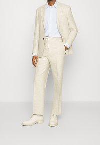 Beige linnen pak met op maat gemaakte blazer en broek met trekkoord, gecombineerd met een lichtblauw overhemd met stippen en crèmekleurige schoenen.