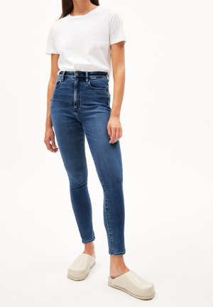 Femme portant un jean skinny bleu taille haute, un t-shirt blanc à manches courtes et des chaussures beiges oversize à enfiler, debout devant un fond blanc.