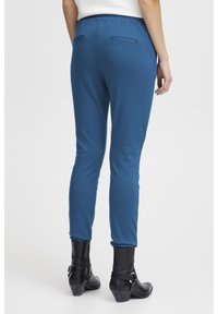 Blå stretchbyxor med elastisk midja, två bakfickor och en figurnära design vid anklarna, tillsammans med svarta ankelstövlar.