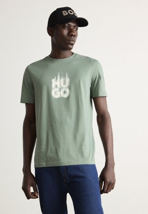 Camiseta de algodón verde claro con mangas cortas que presenta un estampado blanco "HU GO". Combinada con pantalones de mezclilla azules y una gorra negra etiquetada como "BOSS".