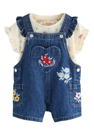Niño con vestido vaquero con bordado floral, calcetines amarillos y camisa con flores, de pie cerca de decoraciones florales pintadas.