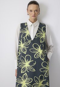 Blomstret kjole i mørkt stof med store gule blomster. V-hals design, båret over en hvid skjorte med krave, glat tekstur.