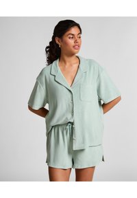 Vrouw met zwart haar in een lichtgroen overhemd met korte mouwen en bijpassende shorts, staand met haar handen achter haar rug en kijkend naar rechts.