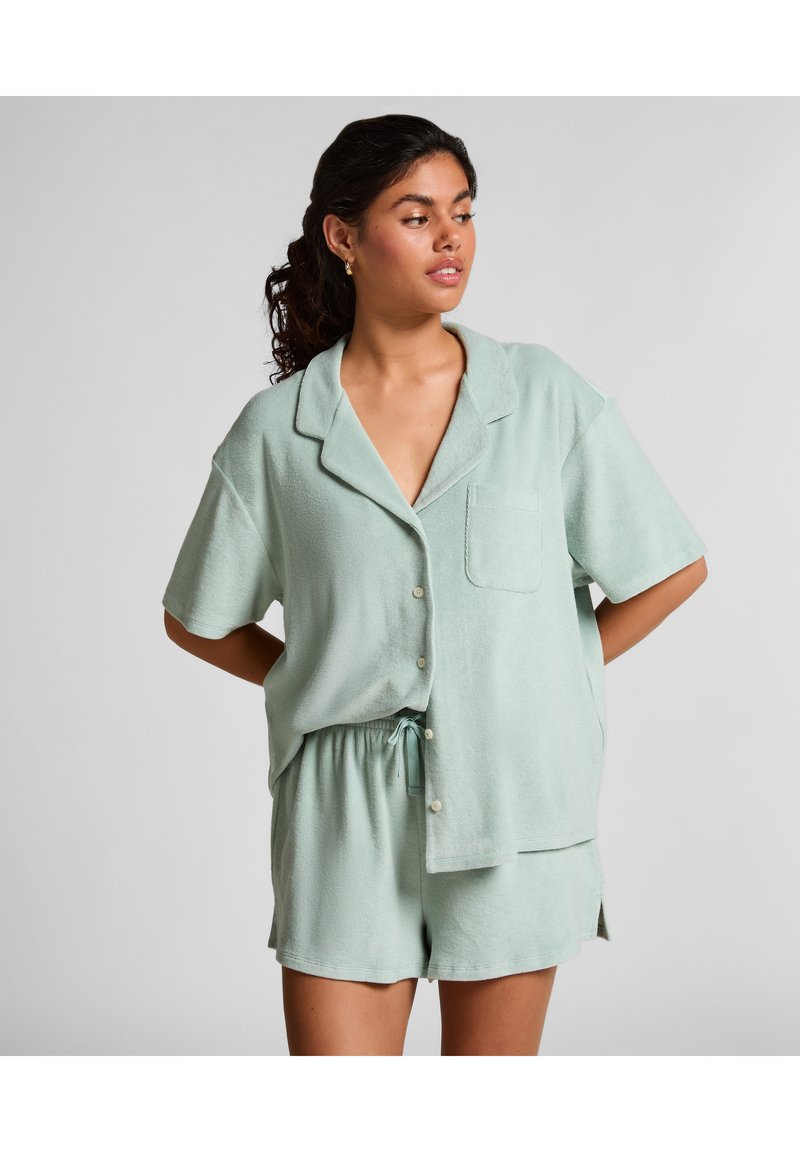 Vrouw met zwart haar in een lichtgroen overhemd met korte mouwen en bijpassende shorts, staand met haar handen achter haar rug en kijkend naar rechts.