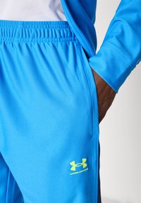 Modre športne hlače s spodnjim vezanjem, narejene iz gladkega materiala. Na levi stegni ima svetlo rumen logotip Under Armour.
