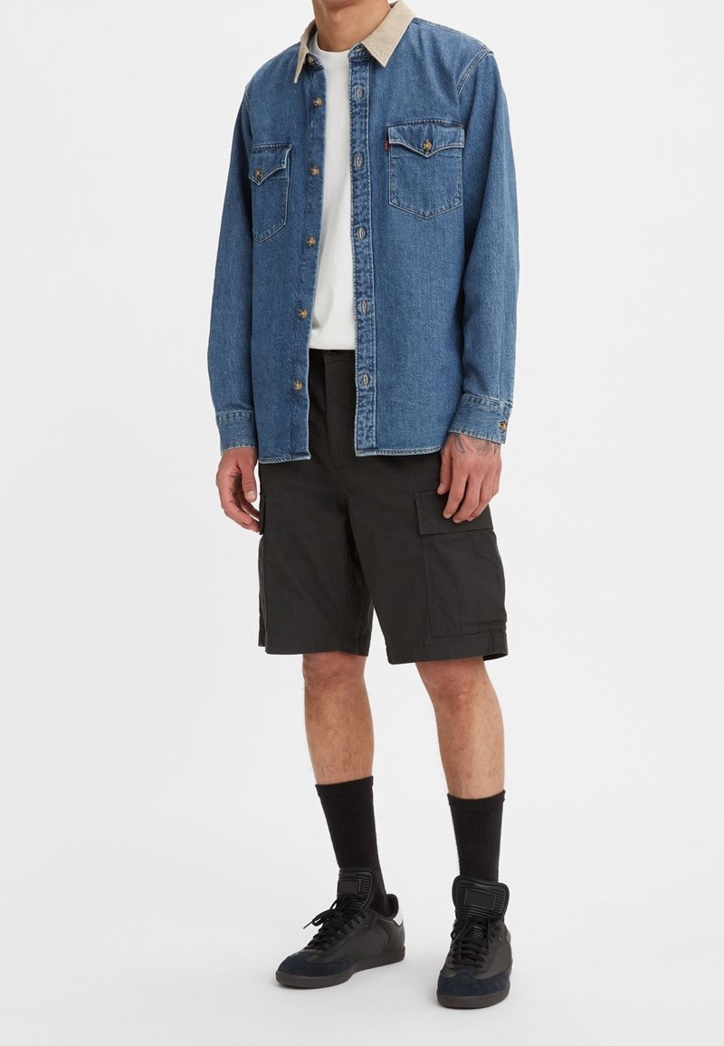 Chemise en denim avec un col beige, deux poches poitrine et une fermeture à boutons, associée à un short cargo noir et des baskets noires.