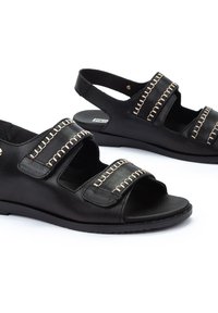Las sandalias de cuero negro cuentan con un diseño de punta abierta, dos cintas anchas con costuras doradas, y un tacón bajo en cuña para mayor comodidad.
