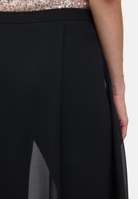 Jupe noire au design fluide, fabriquée en tissu léger, avec une texture lisse et un éclat subtil. Une fente sur le côté ajoute du détail.