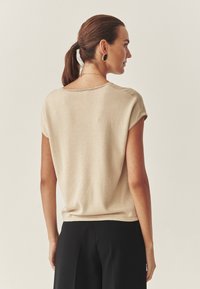 Beige lyhythihainen paita, jossa on sileä pinta ja rento istuvuus, yhdistettynä mustiin housuihin, takaviisto. Yksinkertainen, pyöreä kaula-aukko.