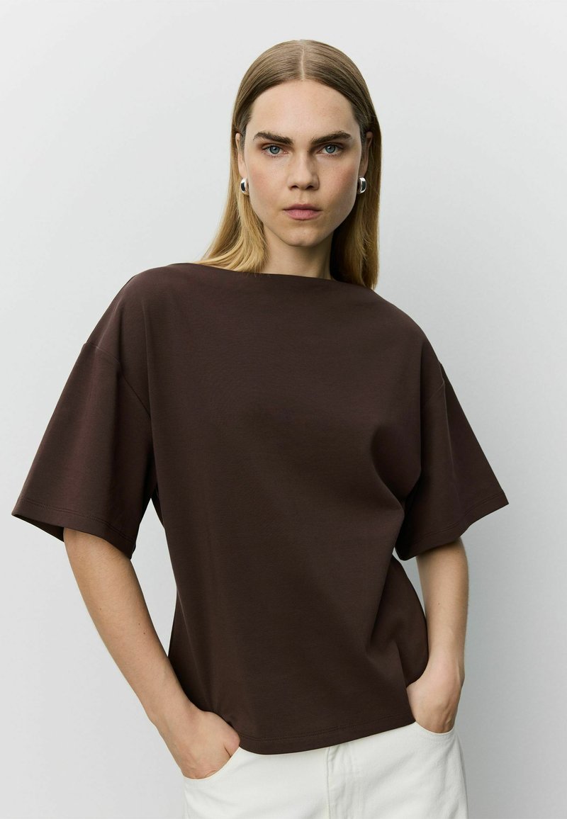 Femme aux cheveux blonds raides, portant des boucles d'oreilles argentées en créoles, un haut ample à manches courtes marron foncé, et un pantalon blanc avec les mains dans les poches.