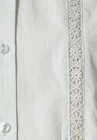 Biała bawełniana koszula z guzikowym zapięciem i ozdobnym, haftowanym panelem z przodu. Gładka tekstura z delikatnym połyskiem.