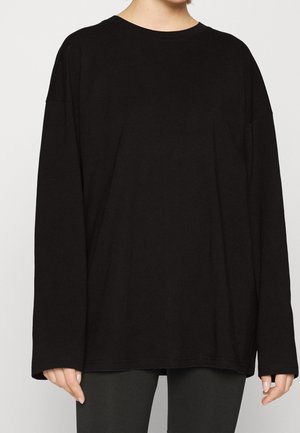 Personne portant une chemise noire ample à manches longues avec un col rond, associée à un pantalon foncé, sur un fond uni.